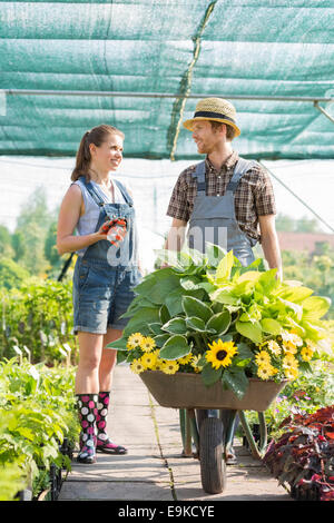 Smiling gardeners discussing while pushing plants in wheelbarrow at greenhouse Stock Photo