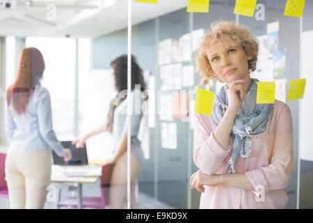 Thoughtful businesswoman reading sticky notes on glass with colleagues in background