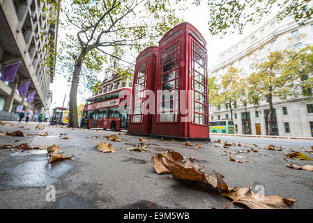 LONDON, United Kingdom — Autumn leaves scatter the footpath on The Strand in central London, framing a quintessential British scene with a pair of iconic red telephone boxes and a red double-decker bus in the background. This image captures the charm of London in fall, showcasing both its natural beauty and iconic urban elements. Stock Photo