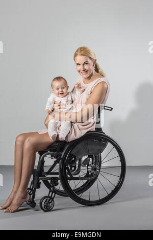 Full length portrait of happy woman with baby girl in wheelchair against gray background Stock Photo