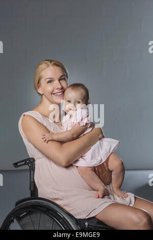 Portrait of happy woman with baby girl in wheelchair against gray wall Stock Photo