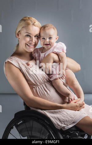 Portrait of happy mother with baby girl in wheelchair against gray wall Stock Photo