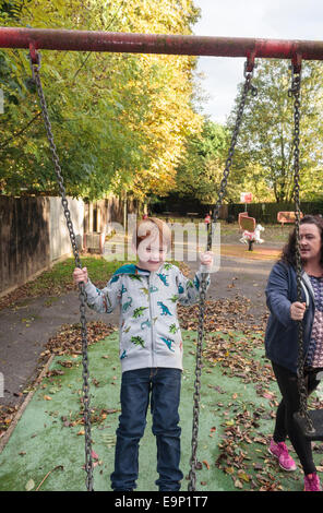 A five-year old boy standing on a swing in a park, being watched by his mother. Stock Photo