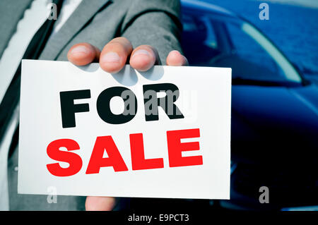 man in suit holding a signboard with the text for sale written in it, with a black car in the background Stock Photo