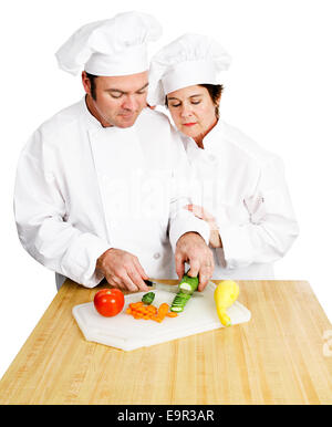 Female chef watches her male student chop vegetables on a cutting board in cooking class.  Both are in full chef's whites.  Isol Stock Photo