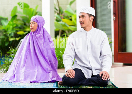 Asian Muslim couple, man and woman, praying at home sitting on prayer carpet in their house in front of the tropical garden Stock Photo