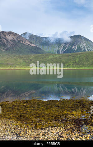 Small white solitary crofters cottage nestling below mountain range reflected in waters of the loch on Isle of Skye in the Highl Stock Photo