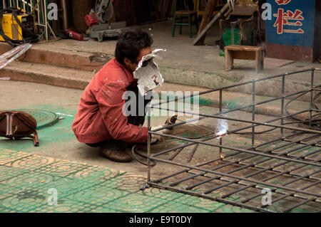 A welder crouches over a metal gate frame, face shielded by newspaper mask, Kaili, Guizhou Province, China Stock Photo