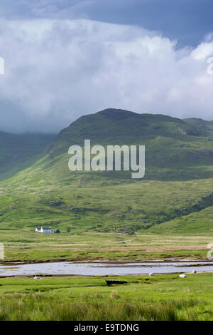 Small white solitary crofters cottage nestling below mountain range by loch on Isle of Mull in the Inner Hebrides and Western Is Stock Photo