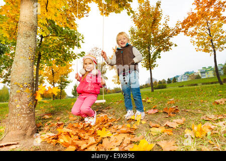 Girl sitting on swings and blond boy standing near Stock Photo