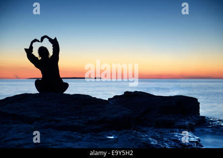 License and prints at MaximImages.com - Silhouette of a woman sitting an a shore at sunset her hands raised in a shape of a flower above her head