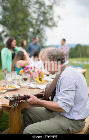 Mature man playing guitar while friends and family talk together after meal, outdoors Stock Photo