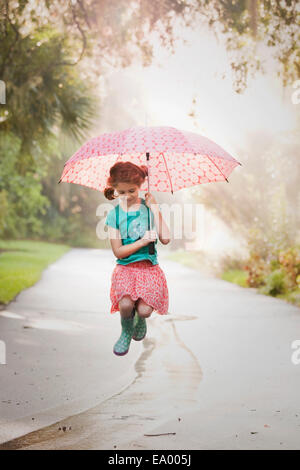 Girl holding up umbrella and jumping puddles on street Stock Photo