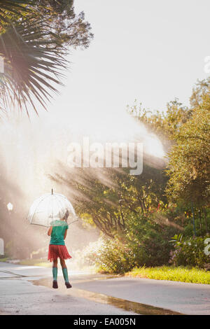 Rear view of girl holding up umbrella and jumping puddles on street Stock Photo