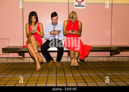 Three young adult friends texting on smartphones whilst sitting in subway station Stock Photo