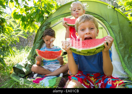 Three children eating large watermelon slices in garden tent Stock Photo