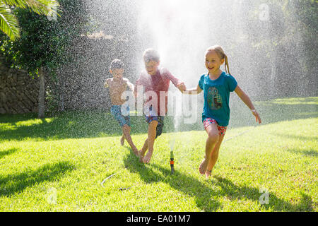 Three children in garden running through water sprinkler Stock Photo