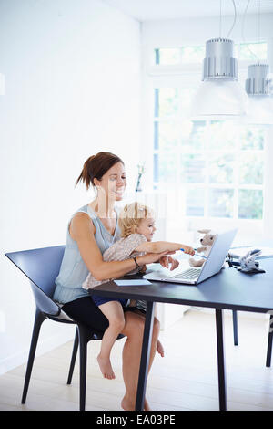 Mid adult woman using laptop with toddler daughter on her lap Stock Photo