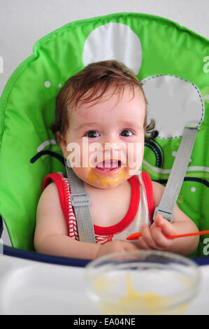 Smiling messy baby sitting in green high chair holding spoon Stock Photo