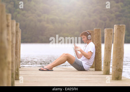 Young man wearing headphones and using mp3 player on jetty over lake