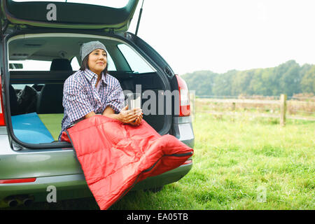 Woman sitting at rear of car with legs tucked in sleeping bag Stock Photo