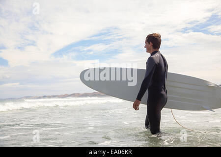 Young man carrying surfboard, walking out to sea