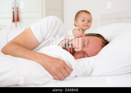 Father asleep in bed, baby daughter watching Stock Photo