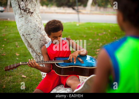 Two brothers sitting on park tree branch playing acoustic guitar Stock Photo
