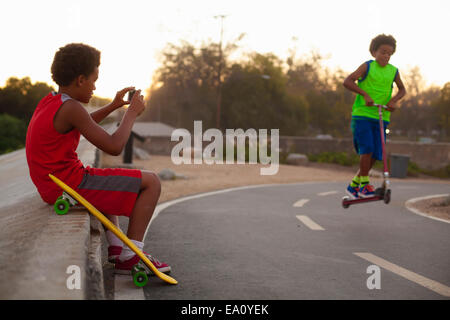 Boy photographing brother doing push scooter jump on road Stock Photo