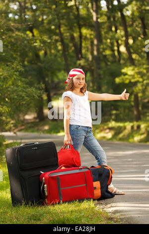 Young hitch-hiker girl standing on road side afternoon in forest with bags Stock Photo