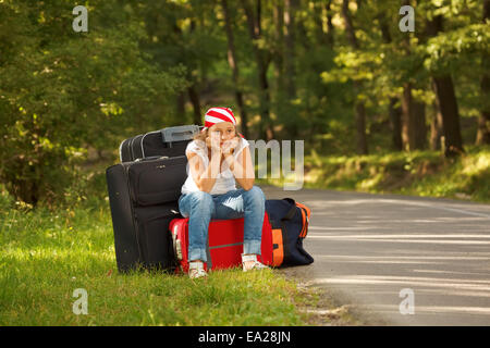 Young hitch-hiker girl standing on road side afternoon with bags Stock Photo