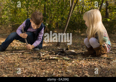 Cute blond little girl playing on the ground with sticks and twigs next to her brother, outdoor in a deciduous forest Stock Photo