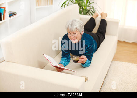 Senior woman laying on sofa, drinking coffee and reading book Stock Photo