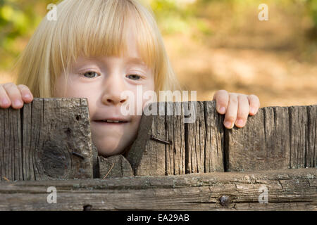 Pretty little blond girl peering over an old rustic wooden fence with a thoughtful expression Stock Photo