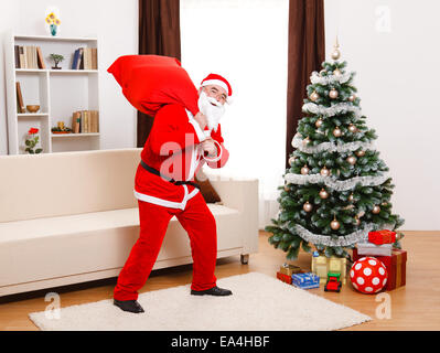 Senior man, wearing Santa Claus uniform, in front of Christmas Tree, walking away with full bag on the back