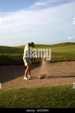 A professional woman golfer hitting his ball out of a bunker with the sand and ball in mid-air