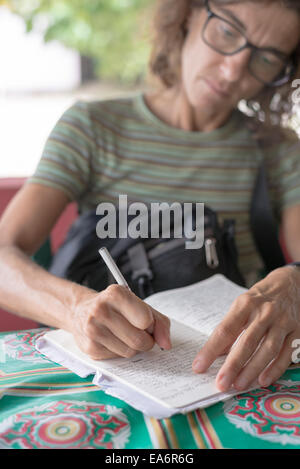 Adult woman with glasses hand writing on notebook with pencil. Selective focus, outdoor location, low contrast, desaturated. Stock Photo