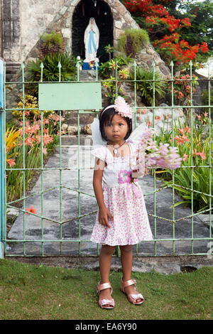 Cute young Filipino girl carries flowers and is dressed in pink for Sunday mass. Stands in front of garden gate.