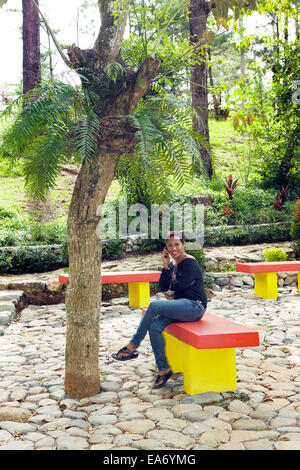 Filipino Asian woman sitting on a park bench outside using her smartphone in Baguio, Philippines. Stock Photo