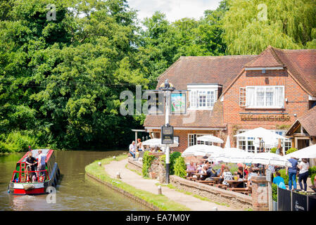 London, UK - July 4, 2014: Narrow boat passes English pub in summer with people sitting outside under umbrellas eating Stock Photo