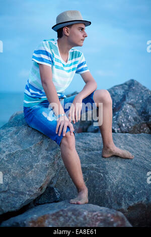 Profile of a handsome teenage boy at sunset sitting on the rocks near seaside Stock Photo