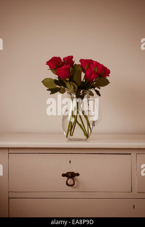 Cerise roses in a glass vase on shabby chic white wooden chest of drawers Stock Photo