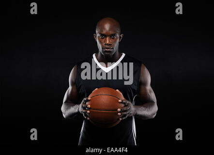 Portrait of muscular young male basketball player in uniform with a basketball posing on black background