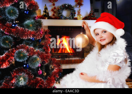 girl in a Santa hat decorating Christmas tree, blazing fireplace on background Stock Photo