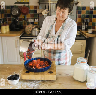 A woman pouring cooked apple and blackberry fruits in to a pie dish. Stock Photo