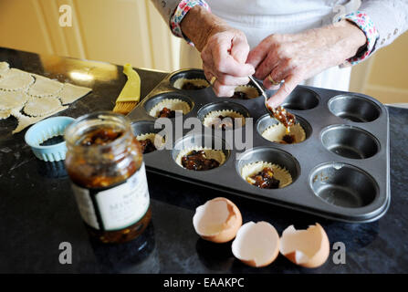 Elderly woman pensioner baking mince pies at home in her kitchen for Christmas . Putting the mincemeat into the pastry shells Stock Photo