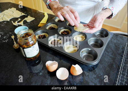 Elderly woman pensioner baking mince pies at home in her kitchen for Christmas . Putting the mincemeat into the pastry shells Stock Photo