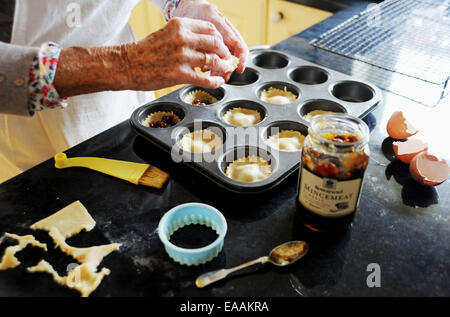 Elderly woman pensioner baking mince pies at home in her kitchen for Christmas . Putting the mincemeat into the pastry shells Stock Photo