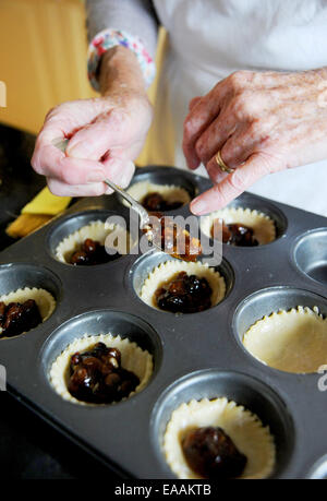 Elderly woman pensioner baking mince pies at home in her kitchen for Christmas . Putting the mincemeat into the pastry shells Stock Photo