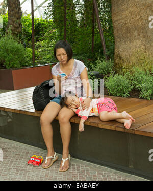 Young Asian woman seated on wooden bench and using mobile phone while young daughter sleeps beside her with head on mother's lap Stock Photo